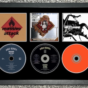 Puede incluir: Obra de arte enmarcada con tres portadas de álbumes y tres CD de Massive Attack y Wild Bunch. Las portadas de los álbumes incluyen "Mezzanine" y "Protection". Los CD son azules, negros y naranjas. El marco es negro.