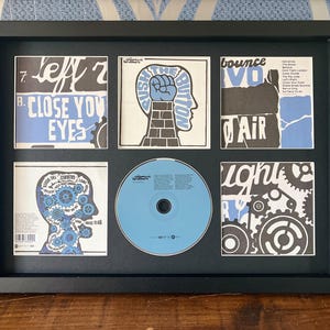 以下が含まれることがあります： CDと6つのアルバムアートの正方形が入った額装されたアートワーク。アートは青、黒、白の色を使用しています。「Close Your Eyes」と「Push the Button」の文字が含まれています。CDは水色です。