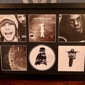 May include: Framed Jamiroquai album art and images. The black frame contains six images, including album covers and band member portraits. The central image is a CD. The overall color scheme is black and white.