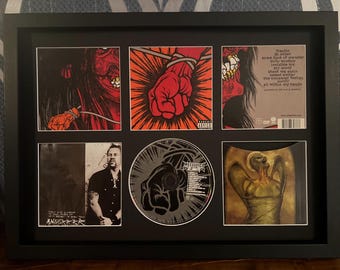 May include: Framed artwork featuring a Metallica album theme. The artwork includes album art, a CD, and a photo of a man. The album art features a red fist, a red face, and a gold figure. The CD has a black fist graphic.