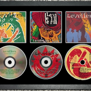 以下が含まれることがあります： LevellersのCDアルバムカバー3枚とCD3枚を額装したコレクション。アルバムアートにはカラフルなイラストとテキストが使用されています。CDは黒いフレームの中にアルバムカバーの下に配置されています。