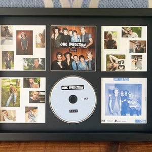 May include: Framed One Direction memorabilia featuring photos and a CD. The black frame holds a CD labeled "FOUR" and multiple small photos of the band members. The album cover is also visible, with the band's name and album title.