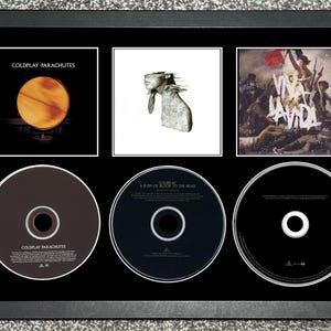 Puede incluir: Obra de arte enmarcada con tres portadas de álbumes y tres CD. La fila superior muestra las portadas de los álbumes de Coldplay: Parachutes, una imagen blanca y Viva la Vida. La fila inferior muestra los CD.