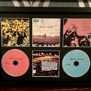 May include: Framed Oasis album art and CDs. The black frame holds six images: three concert photos and three CDs. The CDs are pink and blue, with the text "Time Flies... 1994-2009." The album art features concert scenes.
