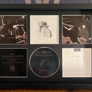 Puede incluir: Exhibición enmarcada de un CD y su arte. El marco negro contiene un CD, arte del álbum y fotos de la banda dispuestas en una cuadrícula. El CD está centrado, rodeado de arte del álbum y fotos de la banda. El álbum se titula "A Rush of Blood to the Head".