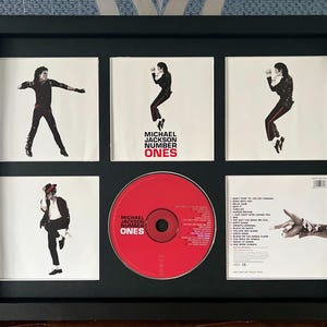 May include: Framed Michael Jackson "Number Ones" CD and album art. The display features a red CD and five album covers with images of Michael Jackson in various poses. The frame is black.