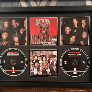 May include: A framed display featuring Death Row Records' Greatest Hits album art and CDs. The black frame contains images of the album cover, artist photos, and two CDs. The album cover is red with "Greatest Hits". The CDs have the number "1".