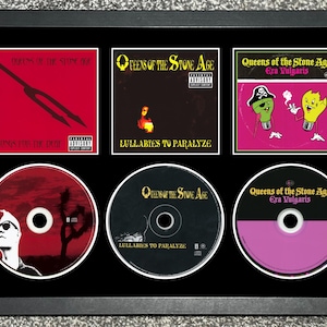May include: Framed display featuring six album covers and CDs by Queens of the Stone Age. The top row displays album art for "Songs for the Deaf", "Lullabies to Paralyze", and "Era Vulgaris". The bottom row shows the corresponding CDs.