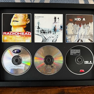 Puede incluir: Una presentación enmarcada en negro con tres portadas de álbumes de Radiohead y tres CD. Las portadas de los álbumes incluyen "The Bends", "OK Computer" y "Kid A". Los CD son visibles debajo. El marco es negro.
