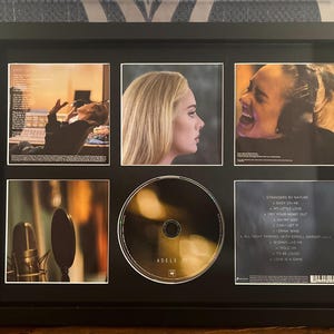 May include: Framed music memorabilia featuring Adele. The black frame holds a CD and several small images, including a profile portrait and a photo of Adele singing. The CD has the artist's name on it. The images include song titles.