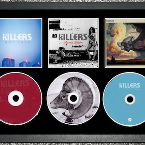 May include: A framed display featuring three album covers and three CDs by The Killers. The album covers include "Hot Fuss," "Sam's Town," and "Imploding the Mirage." The CDs are red, white, and blue, each with the band's name.