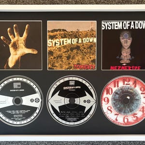 May include: A white-framed display featuring three System of a Down album covers and three corresponding CDs. The top row shows the self-titled album, Toxicity, and Mezmerize covers. The bottom row displays the matching CDs, including the clock-faced disc.