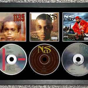 Puede incluir: Obra de arte enmarcada con tres portadas de álbumes y tres CD del artista Nas. Las portadas de los álbumes muestran retratos de Nas, y los CD tienen ilustraciones de álbumes. El marco es negro.