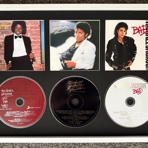 Op de afbeelding: Een ingelijste display met drie Michael Jackson albumhoezen en de bijbehorende cd's: Off the Wall, Thriller en Bad. De witte lijst omringt een zwarte mat die de vierkante albumhoezen boven de ronde schijfjes in een raster plaatst.