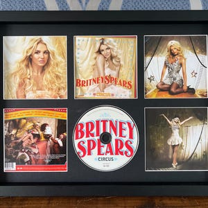 May include: Framed Britney Spears "Circus" CD and album art. The black frame holds six images: three album covers, a CD, and two performance shots. The CD and album covers feature the singer with blonde hair. The album art has the text "Britney Spears Circus".