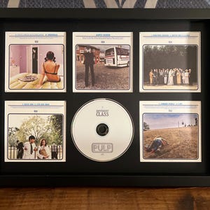 May include: A framed music album display featuring a CD and six album art cards. The CD is white with the text "Pulp Different Class." The album art cards have various images and text. The frame is black.