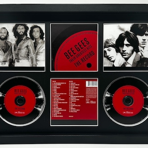 May include: Framed display featuring The Bee Gees album art and CDs. The black frame holds three black and white band photos, a red and black album cover, and two CDs with red labels. The text "BEE GEES" and "THE RECORD" is visible.