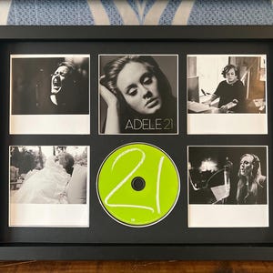 Puede incluir: Exhibición enmarcada en negro con seis imágenes. Cinco fotos en blanco y negro de una cantante y un CD verde con el número "21". El CD está en el centro, rodeado de las fotos. El marco es rectangular.