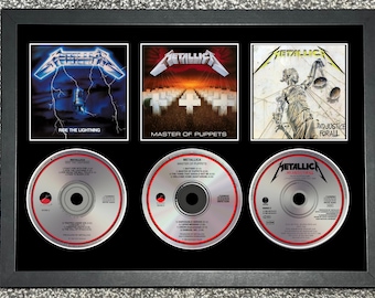 May include: Framed Metallica album art and CDs. The frame contains three album covers: "Ride the Lightning," "Master of Puppets," and "...And Justice for All." Below are three CDs with the Metallica logo. The frame is black.