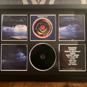 May include: A black framed CD display with six sections. The display includes album art prints and a CD. The album art has a dark, moody aesthetic with blues and purples. The CD has a black surface. Text on the album art includes song titles.