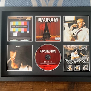 Puede incluir: Obra de arte enmarcada en negro con imágenes y un CD relacionados con el álbum "The Eminem Show" de Eminem. El marco contiene una imagen de pantalla de TV, carátula del álbum y un CD. El CD es rojo con el título del álbum.