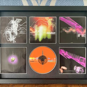Puede incluir: Una presentación enmarcada con un CD y portadas de álbumes de la banda Incubus. El CD es naranja. Las portadas presentan diseños abstractos en blanco, rojo y morado. El marco es negro.