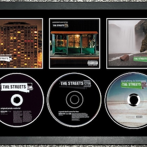 May include: Framed artwork featuring three album covers and three CDs by The Streets. The album covers depict a building at night, a bus stop, and a waterfall. The CDs are black and silver.