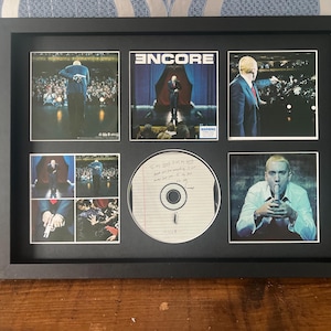 May include: A framed collage featuring images of Eminem. The collage includes album art, concert photos, and a CD. The frame is black, and the images are arranged in a grid. The CD has handwritten text.