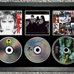 Könnte beinhalten: Gerahmte Präsentation mit drei U2-Album-CDs und drei Albumcover-Bildern. Die CDs sind mit "U2 War", "U2 The Joshua Tree" und einer dritten CD mit Graffiti-Kunst beschriftet. Die Albumcover enthalten Bilder der Band.