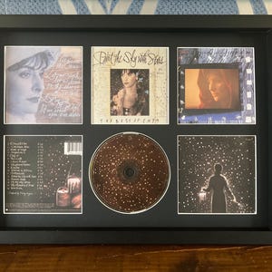 以下が含まれることがあります： EnyaのアルバムアートとCDが額装されています。黒いフレームには、アルバムカバーとCDを含む6つの画像が含まれています。アルバムアートには肖像画と「Paint the Sky with Stars」というテキストが描かれています。CDは茶色で白い点があります。