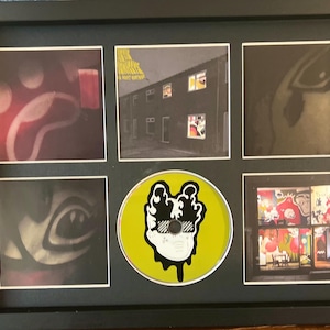 May include: Framed artwork featuring six images, including a CD. The images include album art and a CD with a black and white graphic on a yellow background. The frame is black.