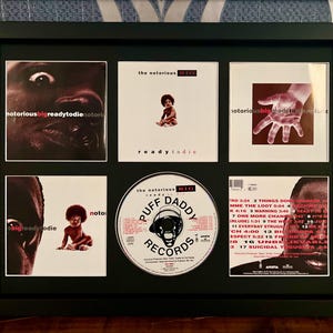 以下が含まれることがあります： The Notorious B.I.G. のアルバムアートとCDを収めた額装作品。 黒いフレームには、アルバムカバーやPuff Daddy Recordsのロゴが入ったCDなど、6つの画像が含まれています。 画像にはテキストとポートレートが含まれています。
