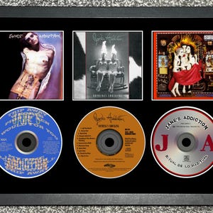 May include: Framed display of Jane's Addiction album covers and CDs. Three album covers are in white frames, and three CDs are below. The CDs are blue, orange, and silver. The album covers feature artistic imagery.