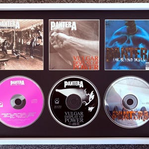 May include: A white framed display featuring three Pantera album covers and their corresponding CDs. The albums are Cowboys from Hell, Vulgar Display of Power, and Far Beyond Driven. The CDs are arranged below their respective album cover art.