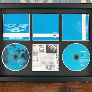 May include: A framed music CD display featuring two CDs with blue and white artwork, three blue album inserts, and a small black and white insert. The frame is black and the background is a patterned blue and white.