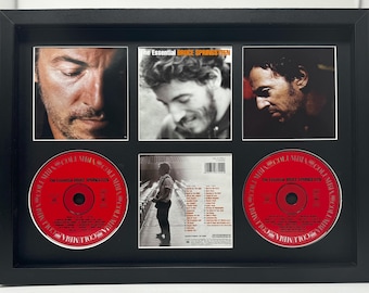 May include: Framed artwork featuring images of Bruce Springsteen, with two red CDs and a CD case. The CDs have the text "The Essential Bruce Springsteen" and the Columbia logo. The frame is black.