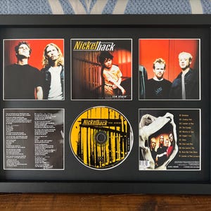 Może przedstawiać: Oprawiona grafika albumu Nickelback i płyta CD. Czarna ramka zawiera sześć obrazów, w tym zdjęcia zespołu, okładki albumów i płytę CD. Płyta CD jest żółto-czarna z nazwą zespołu. Grafika albumu przedstawia zespół i tytuł albumu.