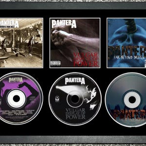 May include: Framed Pantera album art and CDs. The frame holds three album covers: "Cowboys from Hell", "Vulgar Display of Power", and "Far Beyond Driven". Below are three CDs with album art. The frame is black.