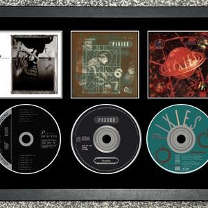 Op de afbeelding: Ingelijste Pixies albumhoezen en cd's. De lijst bevat drie albumhoezen en drie cd's. De albumhoezen zijn in verschillende kleuren, waaronder zwart-wit, bruin en rood. De cd's zijn zwart en blauwgroen.