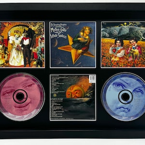 Puede incluir: Obra de arte enmarcada con portadas de álbumes y CD de The Smashing Pumpkins. La obra incluye imágenes de gatos, una mujer en una estrella y niños en un campo. Los CD tienen caras, una rosa y otra azul.