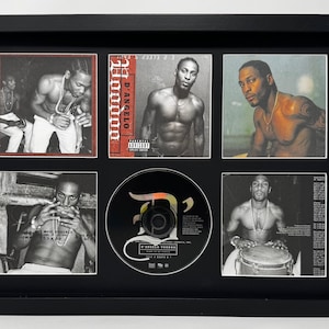 May include: Framed black and white photos and a CD of D'Angelo. The photos show D'Angelo shirtless, with a necklace, and playing instruments. The CD is in the center with the title "Voodoo". The frame is black.