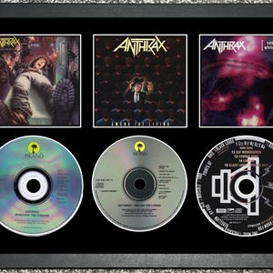 Puede incluir: Enmarcado con arte de álbumes y CD de Anthrax. El marco contiene tres portadas de álbumes y tres CD. Las portadas de los álbumes presentan el logotipo y las ilustraciones de la banda. Los CD están etiquetados con el nombre de la banda y los títulos de los álbumes.