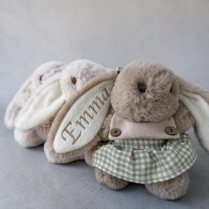 May include: Three plush bunny rabbits in a row. The front bunny is wearing a light pink top and a green and white checkered skirt. The middle bunny has the name "Emma" embroidered on its ear. The bunnies are various shades of brown and beige.