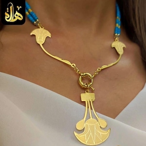 May include: Gold-toned necklace with a pendant featuring a stylized floral design. The necklace has a blue beaded strand and a gold clasp. The pendant is a detailed, symmetrical design. The necklace is worn against a white background.