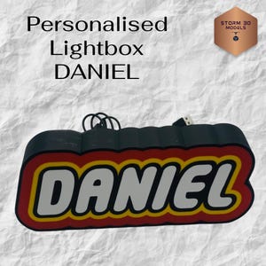 Może przedstawiać: Spersonalizowane pudełko świetlne z imieniem "DANIEL" w białych literach, obramowanych żółtym i czerwonym kolorem, na czarnym tle. Tekst "Personalised Lightbox" znajduje się powyżej. Zawiera kabel USB.