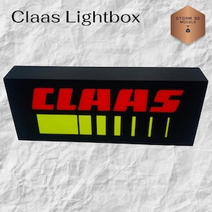 Op de afbeelding: Zwarte rechthoekige lichtbak met het woord "Claas" in het rood en een gele en groene afbeelding. De lichtbak heeft ook de tekst "Claas Lightbox" bovenaan. Een logo staat in de rechterbovenhoek.