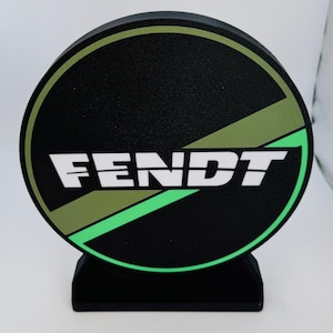 May include: A black and green Fendt logo on a round base. The logo features the word "FENDT" in white, with green and olive green accents. The base is rectangular and black.