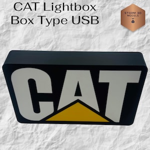 Pode incluir: Caixa de luz CAT preta, tipo caixa USB. A caixa de luz apresenta o logotipo CAT em branco com um triângulo amarelo sobre um fundo preto. O texto "CAT Lightbox Box Type USB" está acima do logotipo.