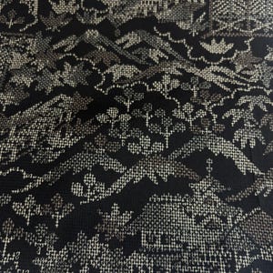 May include: A black fabric featuring a detailed cross-stitch pattern in metallic gold thread. The design depicts a landscape with stylized mountains, trees, and architectural structures, created through a dense grid of small, uniform stitches.