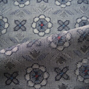 May include: Close-up of a folded fabric with a repeating floral pattern. The design features white, blue, and red accents on a gray background. The fabric has a textured appearance, suggesting a woven material.
